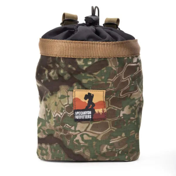 A camouflage drawstring Ape Canyon Outfitters Saddle Pouch Obskura Transitional with a tan trim and a black top. The front features a brown, red, and peach logo patch depicting a silhouette of a person photographing mountains, labeled "APE CANYON OUTFITTERS.