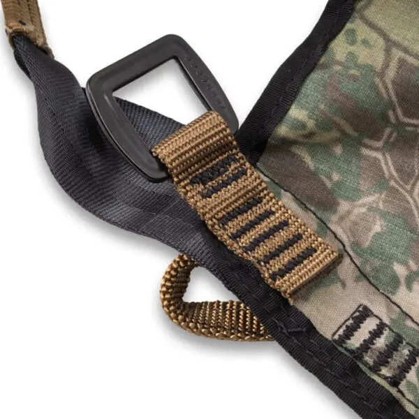 Close-up of a section of a camouflage-patterned fabric with a black trim, featuring a sturdy brown strap threaded through a black plastic rectangular ring. The strap has black stitching for reinforcement. The background is white, highlighting the details of the Ape Canyon Outfitters Pioneer Saddle Obskura Transitional.