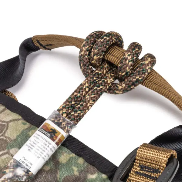 Close-up of a camouflage-patterned Ape Canyon Outfitters Pioneer Saddle Obskura Transitional with brown and black straps. A knotted climbing rope attached to the harness is prominently shown, with black and beige strands. There is a tag with text and a photo of a climber on the rope.