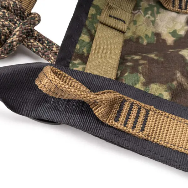 Close-up image of a camouflage-patterned fabric with strong stitched straps in various shades of brown and green. One strap is looped and sewn into the fabric, while a braided rope is partially visible on the left side. The background is white. This is the Ape Canyon Outfitters Pioneer Saddle Obskura Transitional.