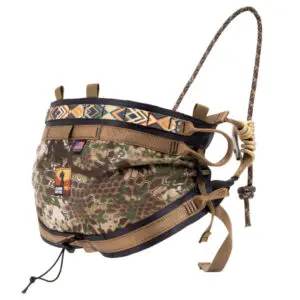 Ape Canyon Outfitters Pioneer Saddle Obskura Transitional: A camouflage-patterned hunting saddle with multiple harness straps and buckles. It features an adjustable rope system and is reinforced with black and tan fabric. The waistband displays a decorative geometric design and a small USA flag patch.