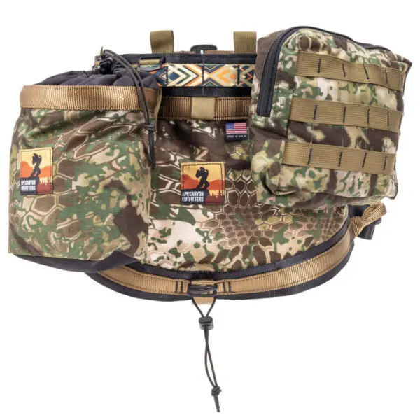 The Ape Canyon Outfitters Pioneer Saddle Obskura Transitional with multiple compartments, including zippered pockets and webbing loops for attaching additional gear. It has a hooped strap for carrying, hunting themed badges, and a small American flag patch.
