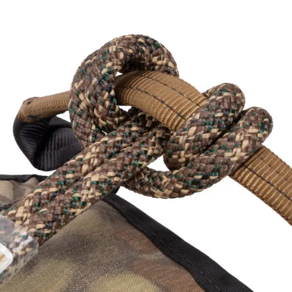 Close-up of an Ape Canyon Outfitters Pioneer Saddle Highlander tied in a knot around a tan nylon strap. The background includes a similarly patterned fabric with black trim. The knot and materials suggest rugged, outdoor or military gear.