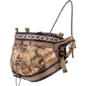 The Ape Canyon Outfitters Pioneer Saddle Highlander is a camouflage-patterned hunting saddle designed for tree stands. It features adjustable straps, buckles, and a padded waist belt with a geometric design. The saddle includes a small American flag patch and an orange silhouette logo.
