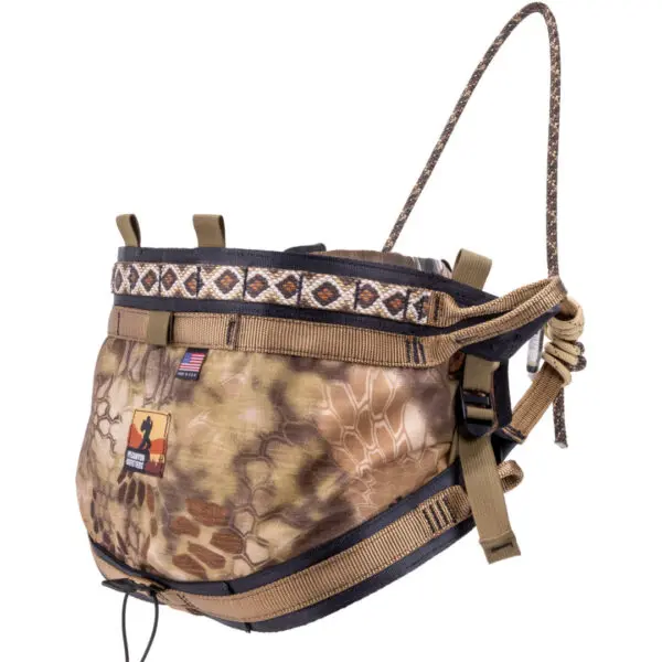 The Ape Canyon Outfitters Pioneer Saddle Highlander is a camouflage-patterned hunting saddle designed for tree stands. It features adjustable straps, buckles, and a padded waist belt with a geometric design. The saddle includes a small American flag patch and an orange silhouette logo.
