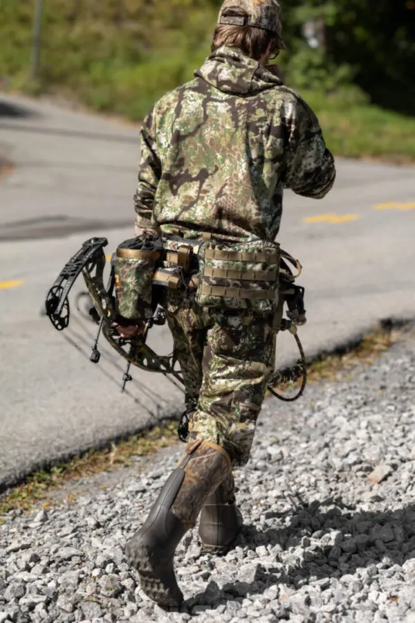 A person dressed in camouflage clothing and rubber boots is walking away on a gravel path. They are carrying an Ape Canyon Outfitters Pioneer Saddle Obskura Transitional and have various gear attached to their belt. The background includes a road and greenery.