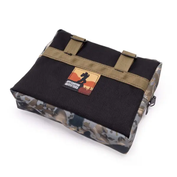 A rectangular black and camouflage bag with a front pocket and a logo featuring a silhouette of a person with "Ape Canyon Outfitters One Stick Saddle Pouch Obskura Skyfall" text. The bag has two olive green straps across the front and a zipper on the side.