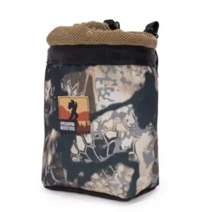A black and tan patterned bag with a mesh top and drawstring closure. The bag features a patch with "Ape Canyon Outfitters Saddle Pouch Obskura Skyfall" and an illustration of Bigfoot in front of a sunset and mountains.