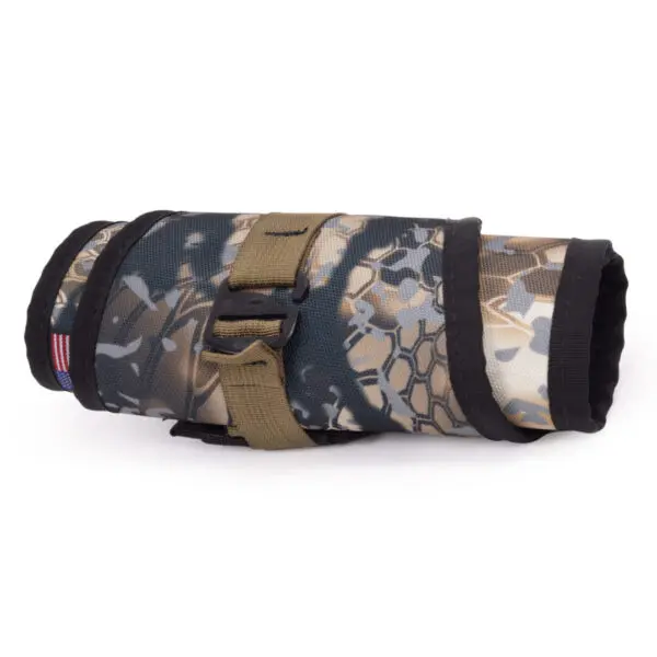 The Ape Canyon Outfitters Lazy Hunter Band Obskura Skyfall is a compact roll-up tool organizer made from camo-patterned fabric with black trim. It has a khaki strap with a buckle securing it, and a small American flag patch on the left side. The organizer is tightly rolled, showing off its durable material and design.