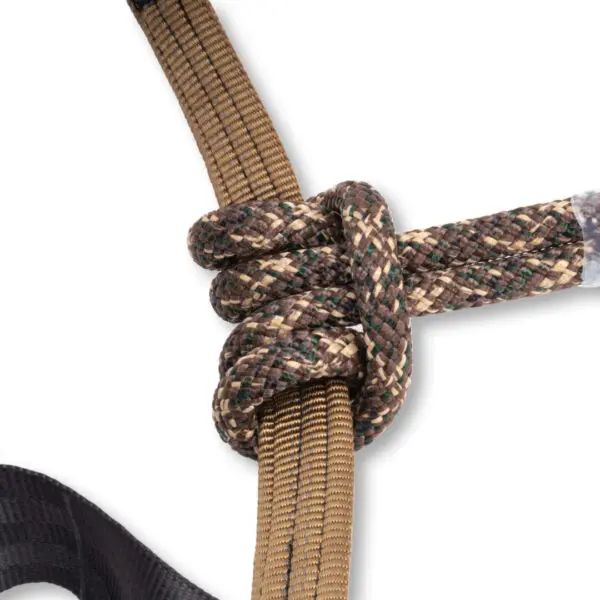 A close-up of an Ape Canyon Outfitters Pioneer Saddle Obskura Skyfall With Cobra Pro Style Buckles tightly tied around another rope using a Prusik knot. The ropes are made of braided material with an outdoor, camouflaged color pattern. The background is plain white.
