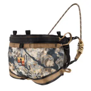 A camo-patterned harness leash designed for outdoor activities, the Ape Canyon Outfitters Pioneer Saddle Obskura Skyfall With Cobra Pro Style Buckles features adjustable straps, black and tan buckles, and a sturdy handle connected at the top by a looped rope. An orange logo with a bear silhouette is on the side.