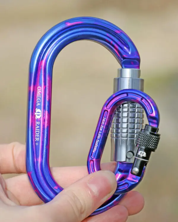 A close-up image of a shiny blue and purple Omega Pacific Micro Raider Aluminum Keylock Screw-Lok held in someone's hand. The carabiner has a silver, textured locking mechanism and intricate details visible, designed for secure connections in climbing and rigging.