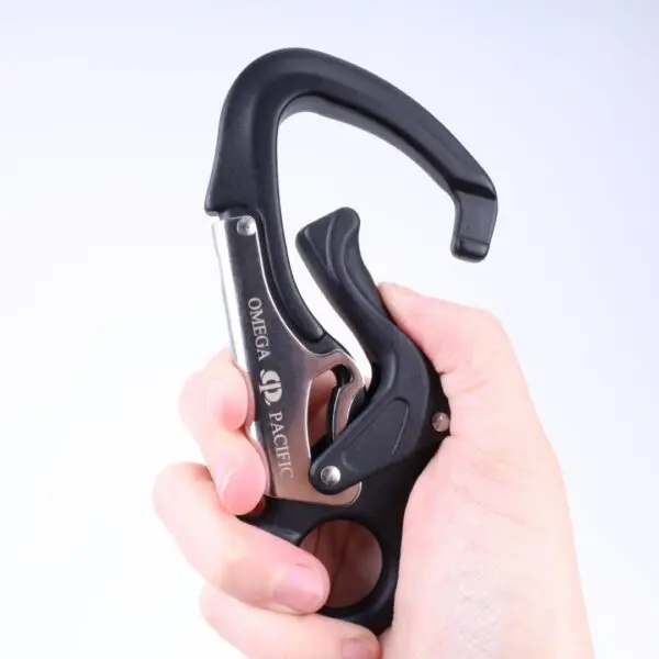 A hand holds a black Omega Pacific Squatch Claw Aluminum Double Action Quik-Lok. The carabiner has a heavy-duty design with a secure, spring-loaded gate that can be pressed open. The brand name "Omega Pacific" and logo are engraved on the metal part of the carabiner.
