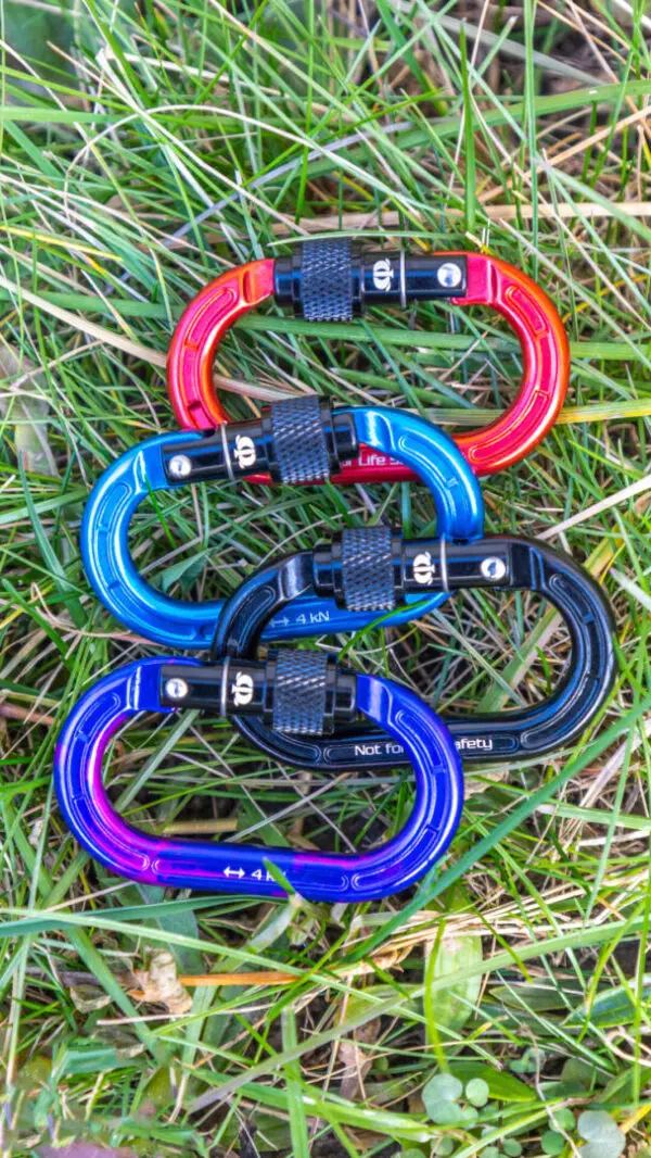 Five Omega Pacific Micro Raider Aluminum Keylock Screw-Lok (red, orange, blue, purple, and black) with screw-lock gates lie on green grass. The carabiners are arranged in a stack, showcasing their vibrant colors and metal construction.