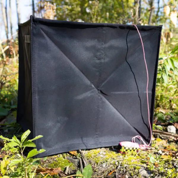 A black RNA collapsible throwline cube with a pink cord stands amongst natural foliage under bright sunlight.