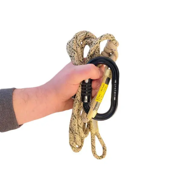 A hand holding an Ape Canyon Outfitters 8mm Oplux Tether System with 3 Stage Quick Lock Carabiner. The person is wearing a grey long-sleeved shirt. The background is white.