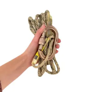 A hand holds an Ape Canyon Outfitters 8mm Oplux Tether System with Screw Lock Carabiner. The rope is beige with black patterns, and the carabiner is metallic with a screw-lock mechanism. The background is plain white.