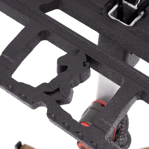 Close-up of a black metal structure with intricate cutouts. In the background, part of an Ape Canyon Outfitters Loaded Prospector One Stick is visible, attached to a bike frame with a red accent on one corner of the pedal. The image highlights the pedal's details and texture.