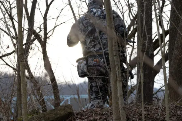 A person dressed in camouflage stands among leafless trees in a forest, facing away from the camera. They appear to be holding an Ape Canyon Outfitters Wood Deluxe Saddle Obskura Skyfall, and the sun is setting in the background.