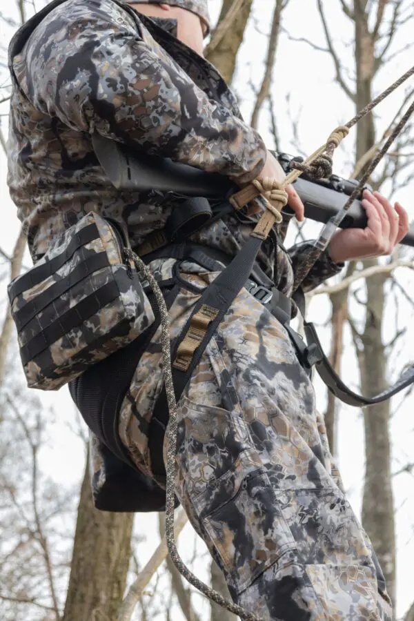 A person in camouflage gear with a rifle is standing outdoors, gripping the firearm's barrel, amidst a background of leafless trees. They have an Ape Canyon Outfitters Wood Deluxe Saddle Obskura Skyfall attached to their waist belt. The scene suggests a hunting or military scenario.