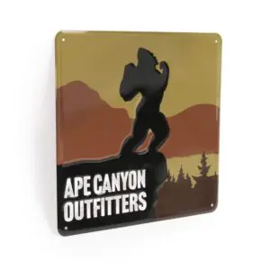 A rectangular metal sign features an illustration of a silhouette of Bigfoot against a backdrop of mountains and pine trees. Below the image, the text reads, "Ape Canyon Outfitters Vintage Metal Sign." The colors are earthy with tones of brown, green, and orange.