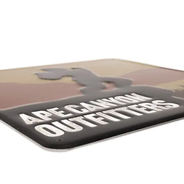 A rectangular sign that reads "Ape Canyon Outfitters Vintage Metal Sign" in bold, white letters with an illustration of a mountain and a stylized Bigfoot silhouette against a gradient background in muted shades of brown and beige.