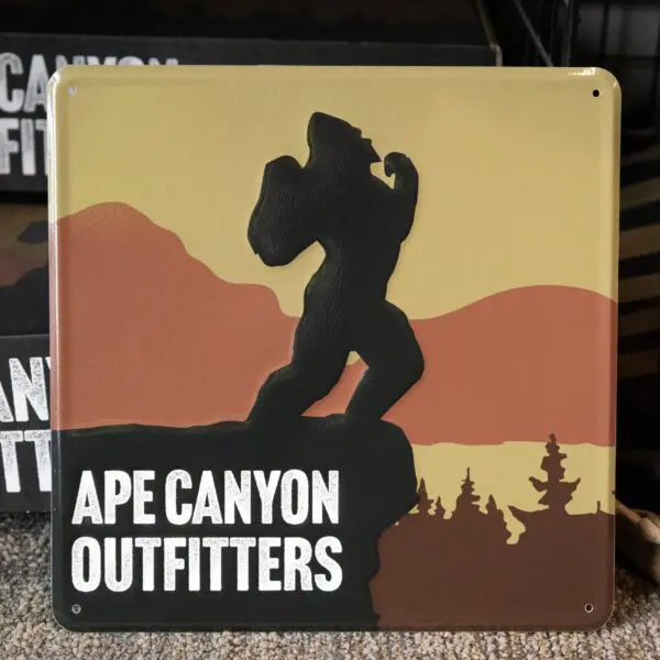A square sign with a silhouette of a Bigfoot-like creature standing on the edge of a cliff against a background of rolling hills and trees. The text "Ape Canyon Outfitters Vintage Metal Sign" appears in bold white letters at the bottom left.