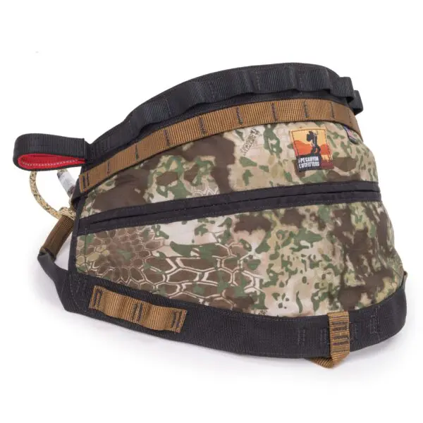 The Ape Canyon Outfitters Wood Deluxe Saddle Obskura Transitional features a camo-patterned tactical dog harness with black, brown, and green accents. It includes a sturdy handle, adjustable straps with buckles, and a patch that reads "IF IT’S CUTE, LIFT IT UP". The harness has a durable design suitable for outdoor activities.