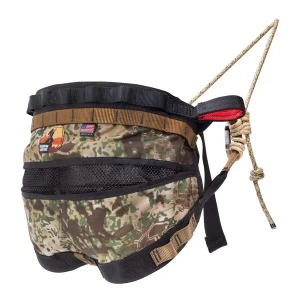 A camouflage-patterned Ape Canyon Outfitters Wood Deluxe Saddle Obskura Transitional with multiple straps, a U.S. flag patch, and a loop for attaching a leash. The harness is designed for rugged outdoor use and features a handle on top for additional control. A rope is also attached to the harness.