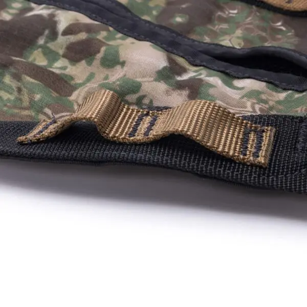 Close-up of a camouflage fabric with a tan-colored strap attached and stitched onto it. The strap has folded segments and is sewn onto black edging. The fabric appears to be part of the Ape Canyon Outfitters Wood Deluxe Saddle Obskura Transitional, laid flat on a white surface.