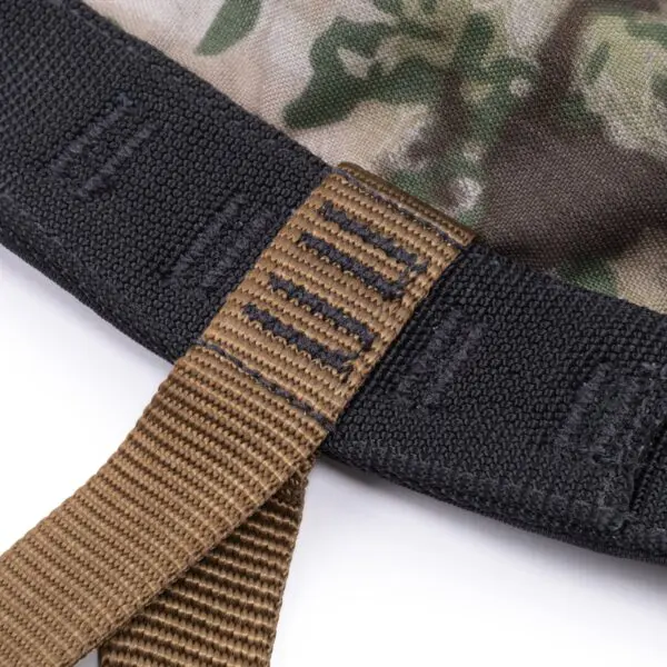 Close-up image of a fabric strap in shades of brown, attached to a piece of material with a camouflage pattern. The strap features reinforced stitching, and the edge of the fabric has black stitching with loop attachments. The background is plain and white. Product: Ape Canyon Outfitters Wood Deluxe Saddle Obskura Transitional