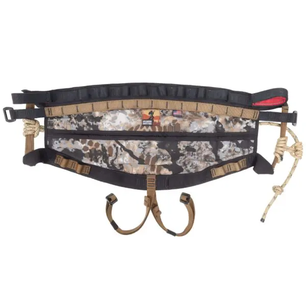 A camouflage-patterned Ape Canyon Outfitters Wood Deluxe Saddle Obskura Skyfall with adjustable straps and buckles. The saddle features padded support, multiple attachment loops, and rope elements, designed for tree hunting.