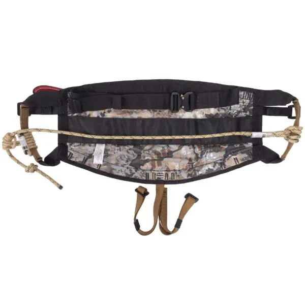 A camouflage-patterned rock climbing harness with black and brown straps is shown, featuring multiple adjustment buckles and loops. The Ape Canyon Outfitters Wood Deluxe Saddle Obskura Skyfall has attached ropes with knots at the ends and leg loops.