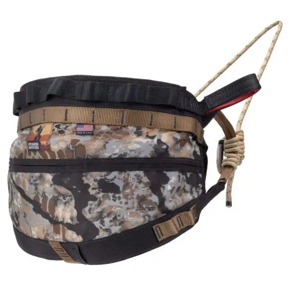 A camouflage-patterned Ape Canyon Outfitters Wood Deluxe Saddle Obskura Skyfall with a black strap and tan accents, equipped with a drawstring closure. The bag has a small American flag patch and another patch on the side. The strap is attached to a knotted beige cord, likely for fastening or carrying.