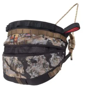 The Ape Canyon Outfitters Wood Deluxe Saddle Obskura Skyfall features a camouflaged bucket organizer with multiple compartments and a drawstring closure. It includes a handle, various webbing, and a small patch with an American flag and a logo. The organizer has a mix of black, brown, and gray tones.