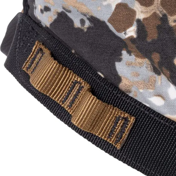 Close-up of a fabric item with a camouflage pattern in black, gray, white, and brown. The fabric has a black edge trim and is fitted with a brown, double-stitched, webbed strap loop from the Ape Canyon Outfitters Wood Deluxe Saddle Obskura Skyfall.