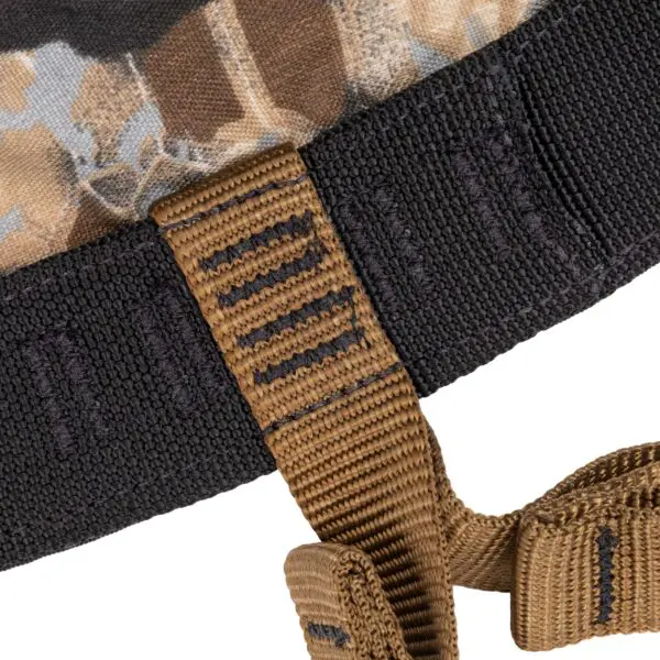 Close-up image of a piece of fabric with a camouflage pattern and a textured black edge. A brown strap with black stitching is looped through a reinforced black band on the fabric. The background is white. The product shown is the Ape Canyon Outfitters Wood Deluxe Saddle Obskura Skyfall.