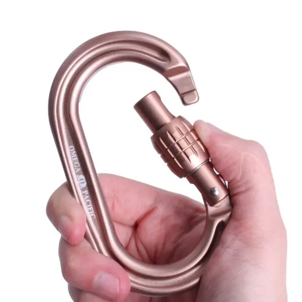 A hand is holding a sand-tan Omega Pacific Raider Tactical Oval Screw-Lok. The carabiner is designed with a locking mechanism to ensure safety and security. It appears sturdy and is intended for use in activities requiring strong and reliable gear, like climbing or rescue operations.