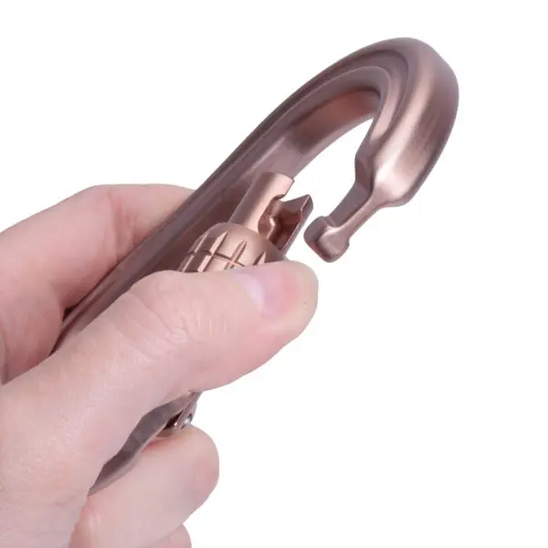 A hand holding an Omega Pacific Raider Tactical Oval Screw-Lok Sand Tan with a screw lock mechanism partially opened. The person's fingers are gripping the carabiner, and the screw lock is visible and textured for a better grip.