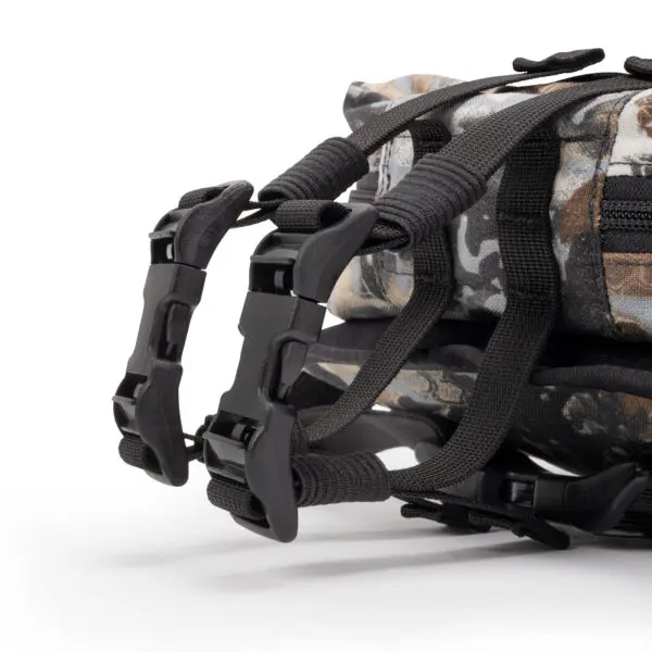 Close-up of two black plastic buckles on an Ape Canyon Outfitters Canyon Quiver Highlander, connected to black straps. The fabric has various shades of gray, green, and brown, mimicking a natural environment. The buckles are fastened securely, ensuring the straps are tightly held in place.