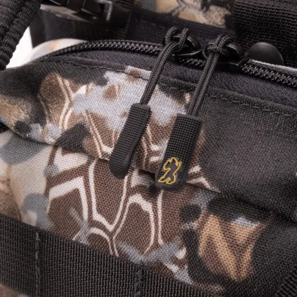 Close-up of the zipper pulls on an Ape Canyon Outfitters Canyon Quiver Highlander with a camouflage pattern. The two black zipper pulls feature a small gold logo of a lion. The bag's fabric has shades of brown, black, and gray forming the camo design, with black stitching and webbing for added detail.