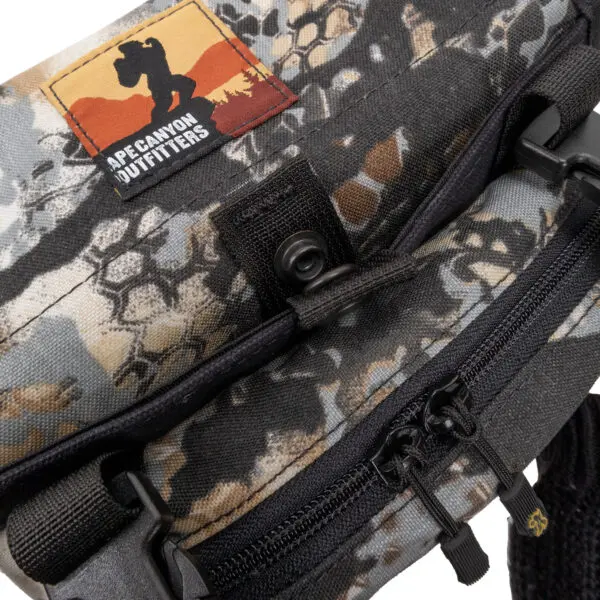 Close-up of a camouflage-patterned bag with a black zipper and a small black buckle. The bag features a rectangular patch with a silhouetted figure holding a camera, labeled "Ape Canyon Outfitters Canyon Quiver Highlander" against a sunset background in the upper left corner.