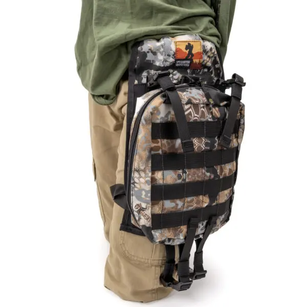 A person wearing khaki pants and a green shirt carries an Ape Canyon Outfitters Canyon Quiver Highlander on their lower back. The backpack features a webbing grid for attachments and has a patch with an orange and black design at the top.