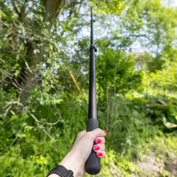 A person with bright pink nail polish holds a JRB Maverick Pole raised in an outdoor wooded area, with greenery and trees in the background. The person is wearing a black wristband on their left wrist.