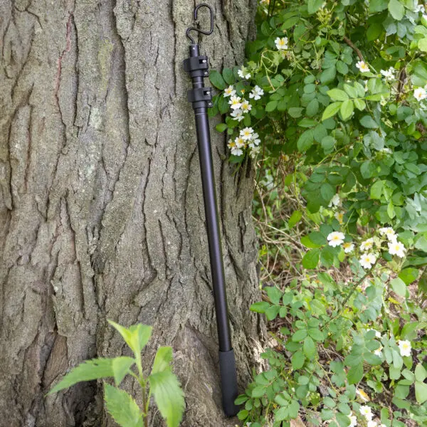 A black JRB Maverick Pole with a foam grip and wrist strap is propped against a tree trunk. In the background, there are green leaves and small white flowers on a bush.