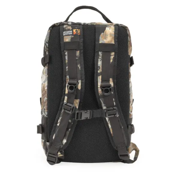 A camo-patterned Ape Canyon Outfitters Conclave Pack Highlander Skyfall with black adjustable straps and a padded back. The bag has a top handle and several loops for attachments. A label on the top of the backpack features a logo and text which is not fully visible in this angle.