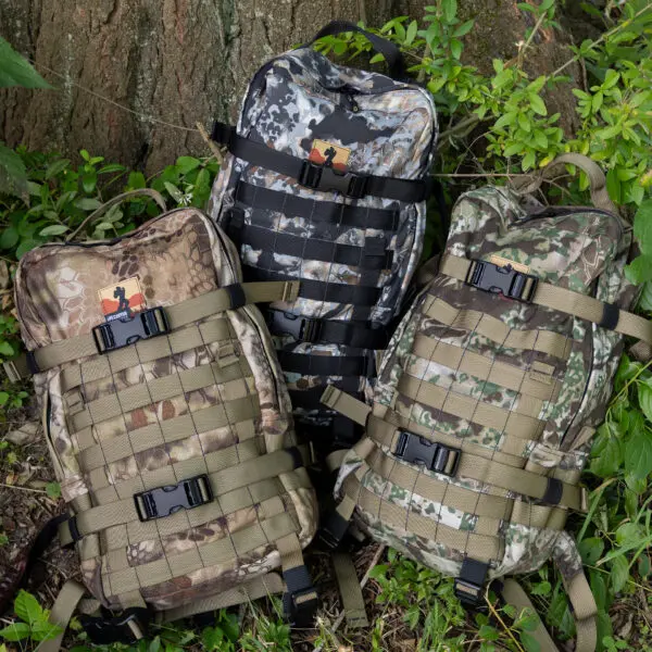 Three Ape Canyon Outfitters Conclave Pack Highlander backpacks are propped against a tree surrounded by greenery. Each backpack features multiple straps and buckles for securing additional items. The patterns include brown, grey, and green camouflages.