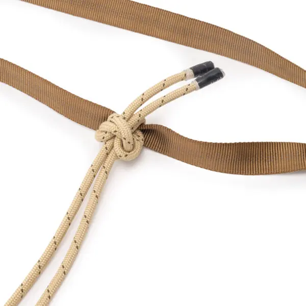 Close-up image showing a tan cord with black tips tied in a secure knot around a brown strap. The strap extends horizontally across the frame, while the cord forms a loop and two loose ends pointing downward. The knot is prominently centered, part of the JRB Garda Hitch Footloop Kit.