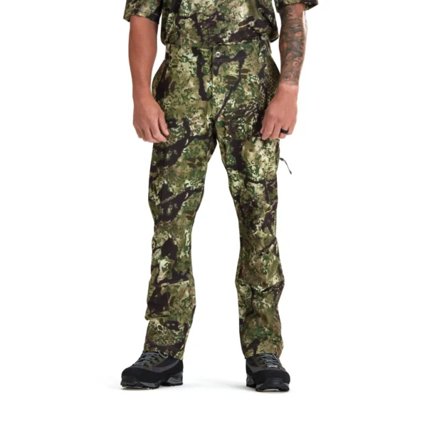 A person is shown from the shoulders down wearing Kryptek Valhalla Pants Obskura Transitional. The person has a tattoo on one forearm and is also wearing black shoes. The white background emphasizes the details of the camo design.