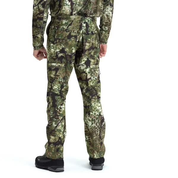A person wearing a full camouflage outfit with long sleeves and Kryptek Valhalla Pants Obskura Transitional is standing with their back to the camera. Their hands are relaxed by their sides, and they are wearing black shoes. The camo pattern features various shades of green and brown.